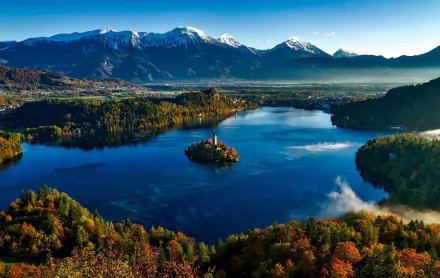 A stunning aerial view of Lake Bled in Slovenia, showcasing the Assumption of Mary Church on an island, surrounded by vibrant autumn foliage and majestic mountains reflecting in the water.