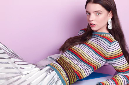 HD desktop wallpaper of American actress and model Anya Taylor-Joy with brunette hair, brown eyes, wearing statement earrings and a colorful striped top against a pink background.