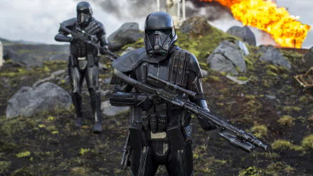 Two Death Troopers wielding blasters stand alert amidst rocky terrain with an explosion in the background. HD desktop wallpaper from the movie Rogue One: A Star Wars Story.