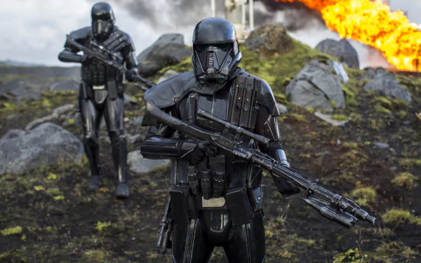 Two Death Troopers wielding blasters stand alert amidst rocky terrain with an explosion in the background. HD desktop wallpaper from the movie Rogue One: A Star Wars Story.