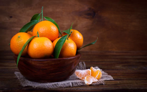 A wooden bowl filled with fresh mandarins with green leaves, displayed on a rustic cloth against a dark wooden background in 4K Ultra HD quality.