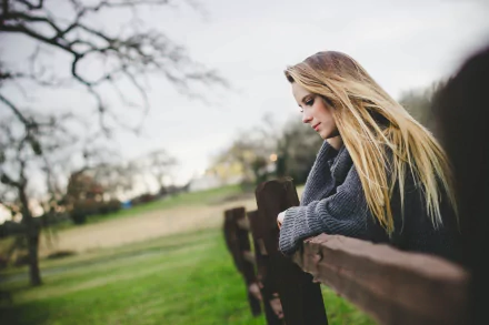 Blonde woman leaning on a wooden fence in soft focus with a moody atmosphere, captured with depth of field for a serene HD desktop wallpaper backdrop.