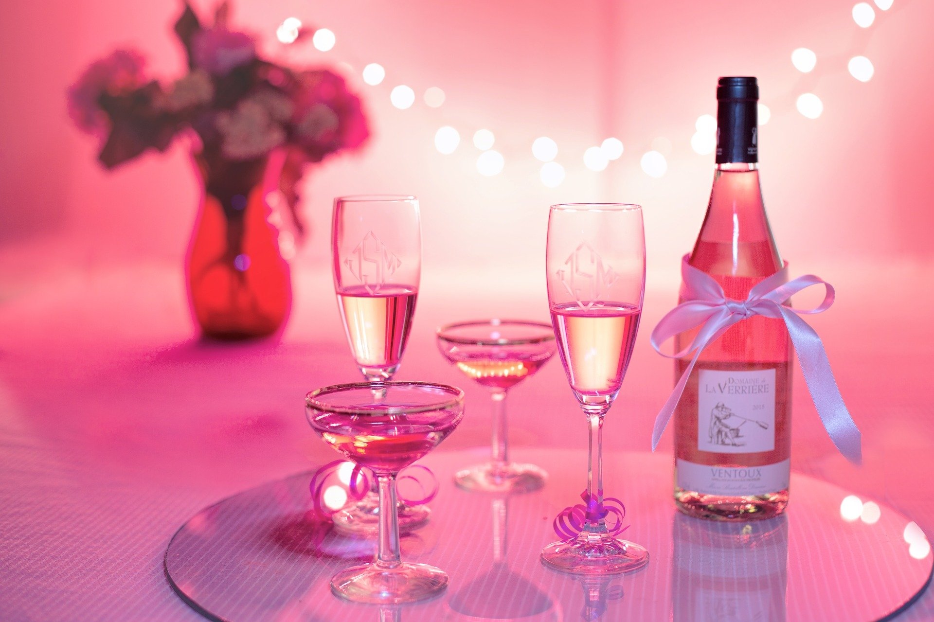 HD still life desktop wallpaper featuring pink champagne glasses and a bottle adorned with a white ribbon, set against a soft pink background with blurred lights and flowers.