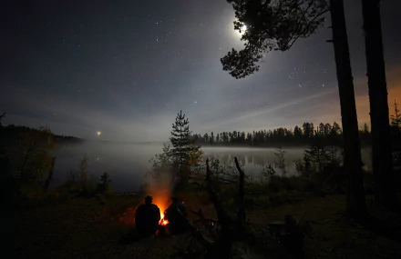 Two people sit by a bonfire near a lake, surrounded by a forest under a starry night sky with the moon shining brightly. This HD desktop wallpaper captures the serene nature of a Russian landscape.