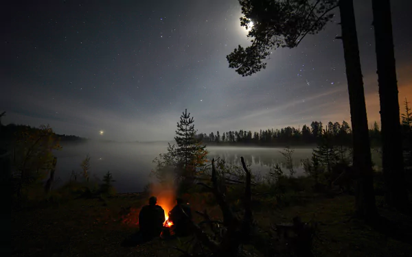 Two people sit by a bonfire near a lake, surrounded by a forest under a starry night sky with the moon shining brightly. This HD desktop wallpaper captures the serene nature of a Russian landscape.