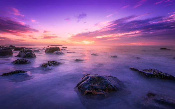HD desktop wallpaper of a purple sunset over the ocean, with boulders scattered in the water and a dramatic sky at dusk on the horizon.