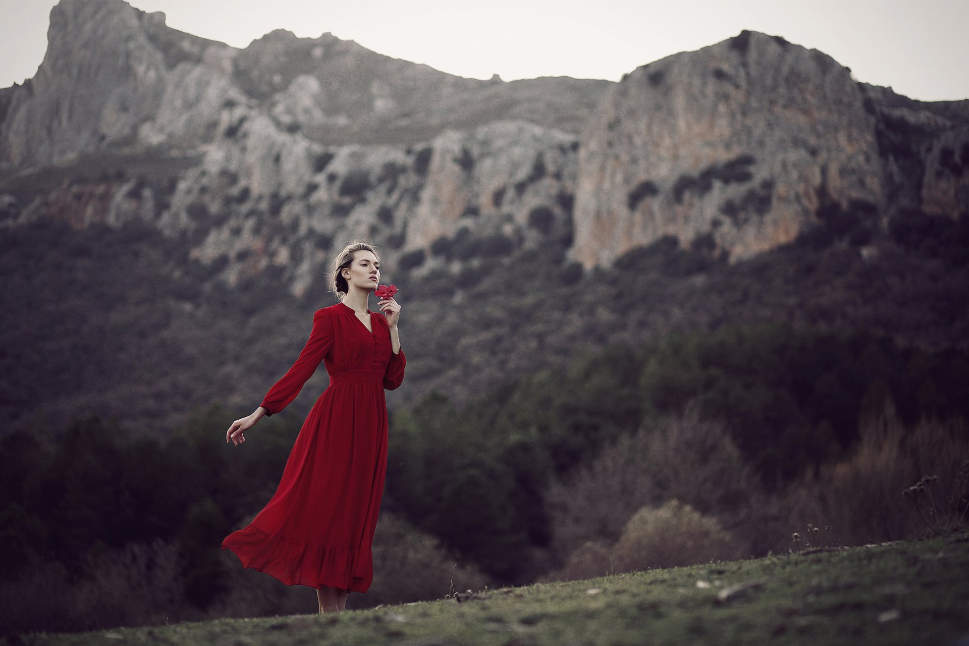 Moody HD desktop wallpaper: a woman model in a flowing red dress stands against a blurred mountain landscape, dramatic depth of field emphasizing her solitary, contemplative pose.
