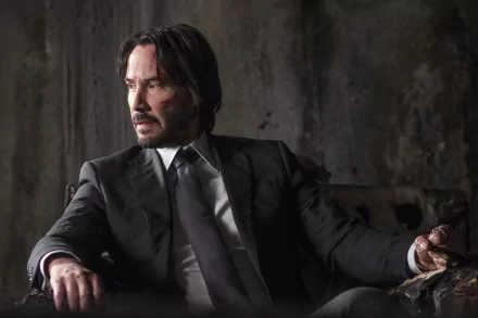 HD desktop wallpaper of Keanu Reeves as John Wick from the movie John Wick: Chapter 2. He is seated in a suit, looking intently to the side with a rugged background.