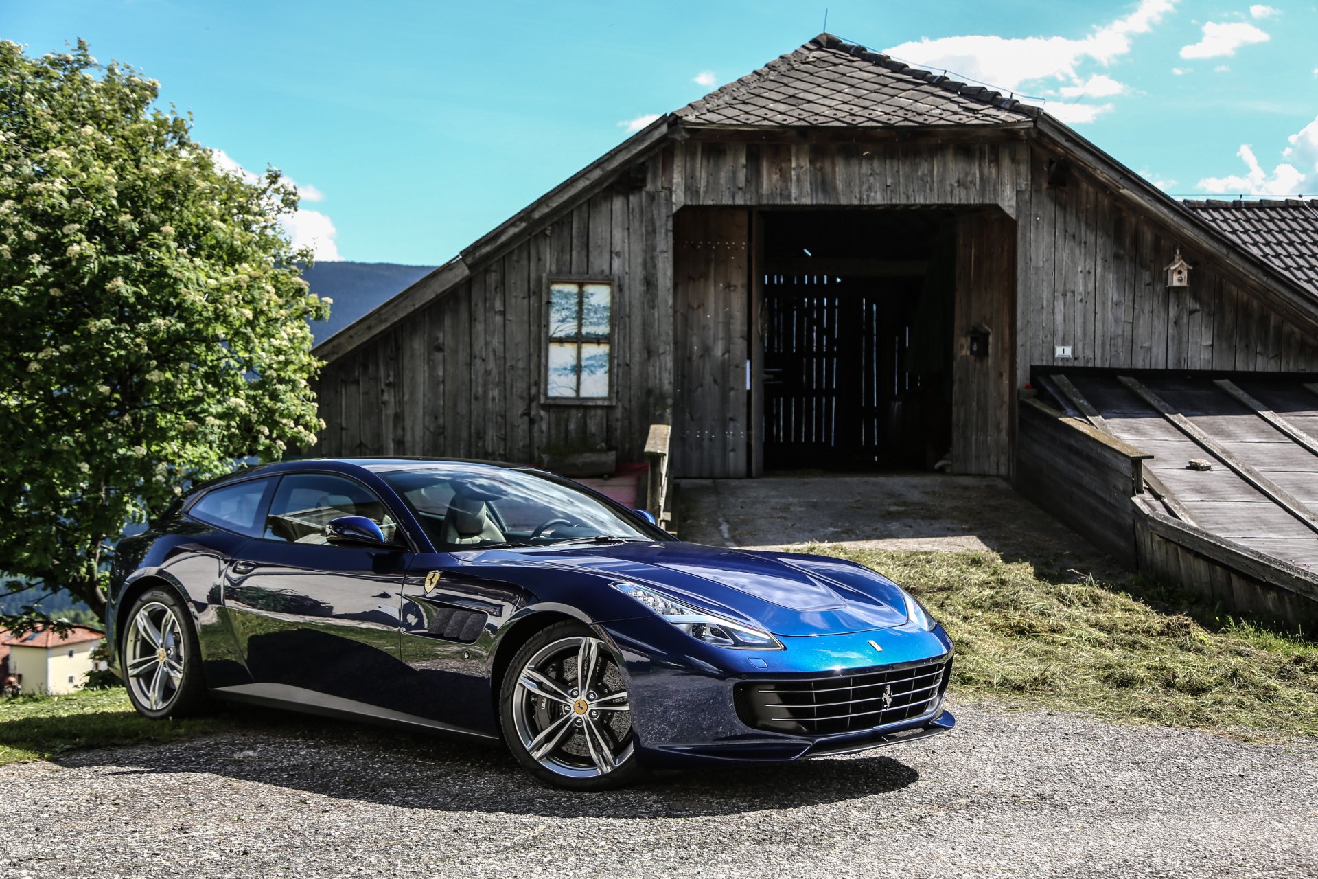 A sleek blue Ferrari GTC4Lusso grand tourer parked in front of a rustic wooden barn, captured in a high-resolution 4K Ultra HD desktop wallpaper.