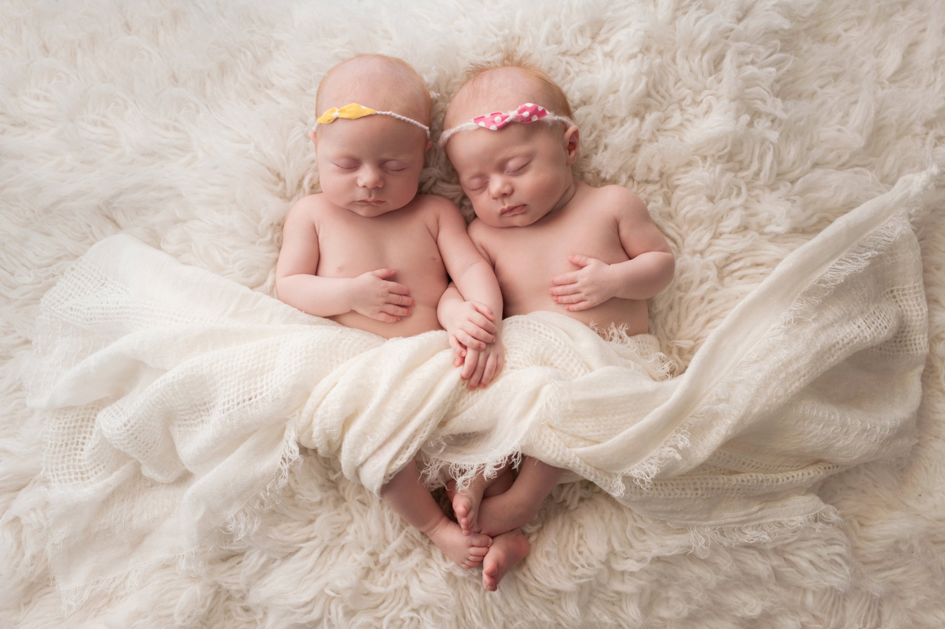 A 4K Ultra HD photo of two sleeping babies with headbands, wrapped together in a soft cream blanket on a fluffy white surface, capturing a cute and serene moment.