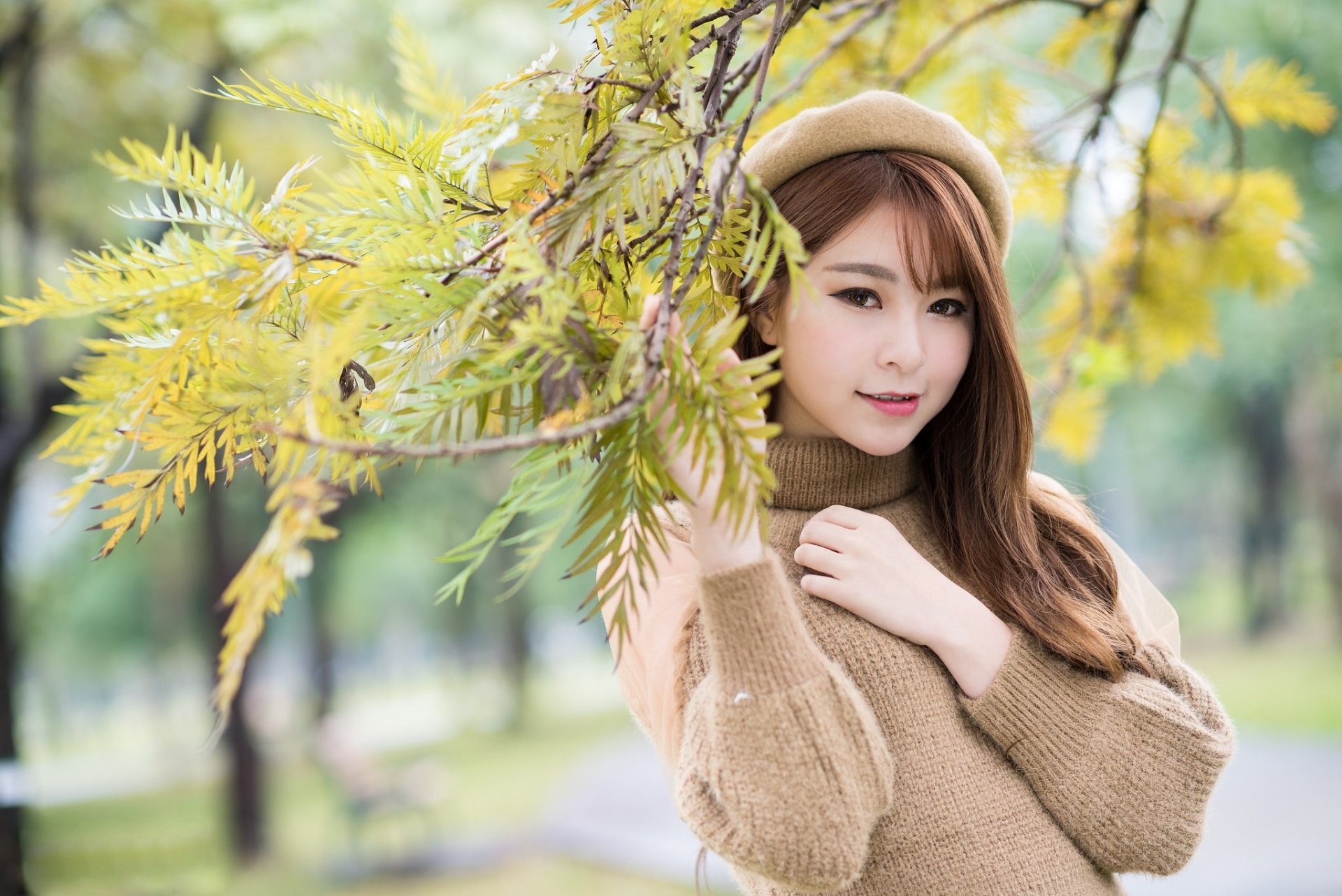 A brunette Asian model with brown eyes and a hat poses outdoors among yellow-green foliage, captured with a beautiful depth of field, making for an elegant HD desktop wallpaper and background.