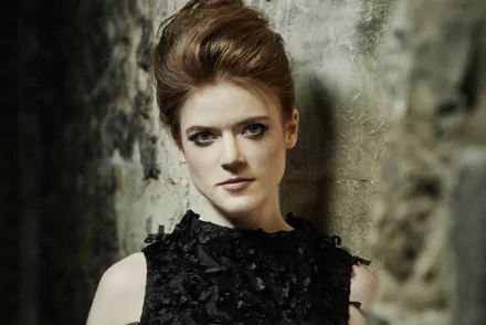 HD desktop wallpaper featuring Rose Leslie, a redhead actress, posing against a textured stone wall in a black textured dress.