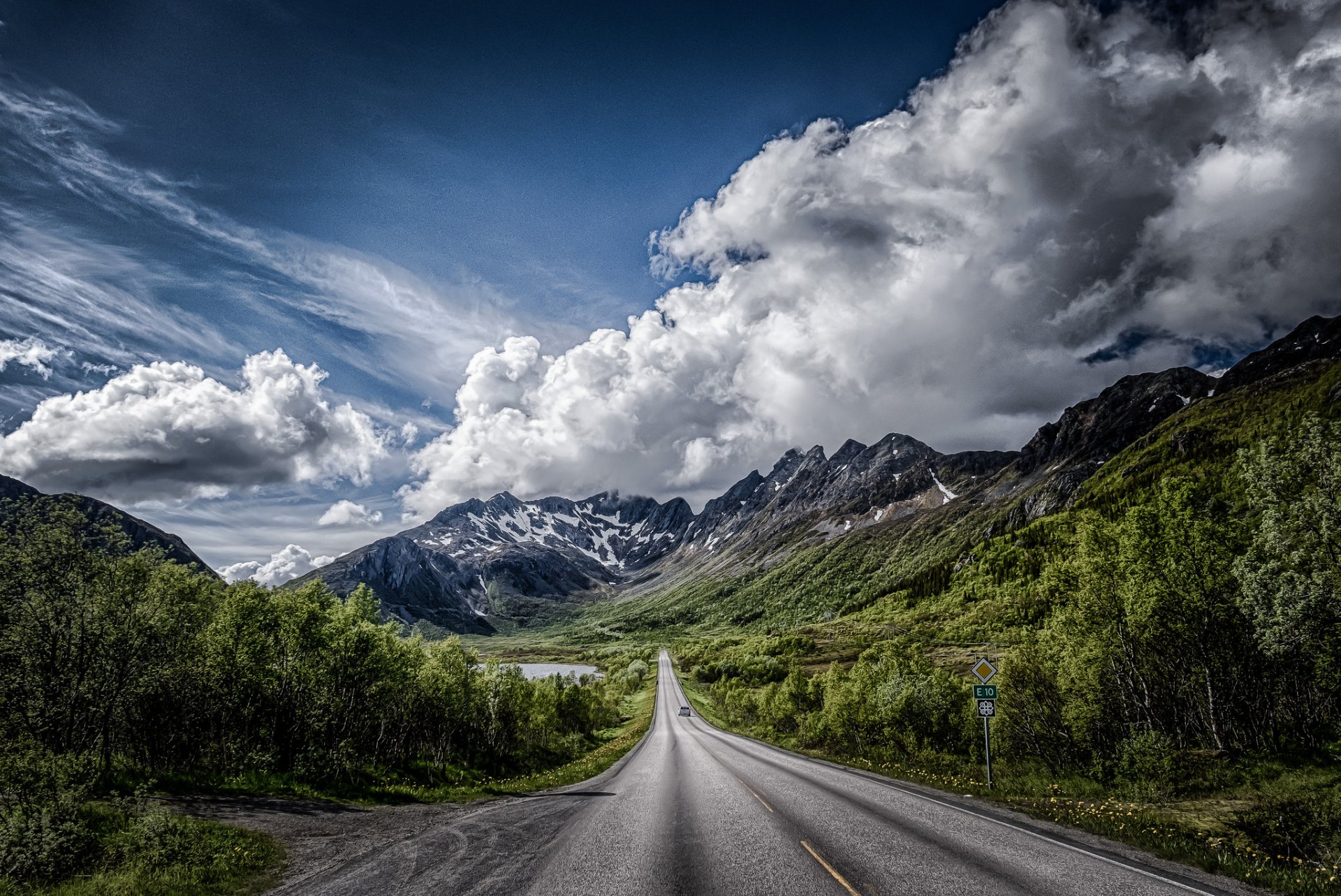 HD desktop wallpaper featuring a road cutting through lush green valleys with towering mountains under a dramatic cloudy sky in a stunning landscape setting.