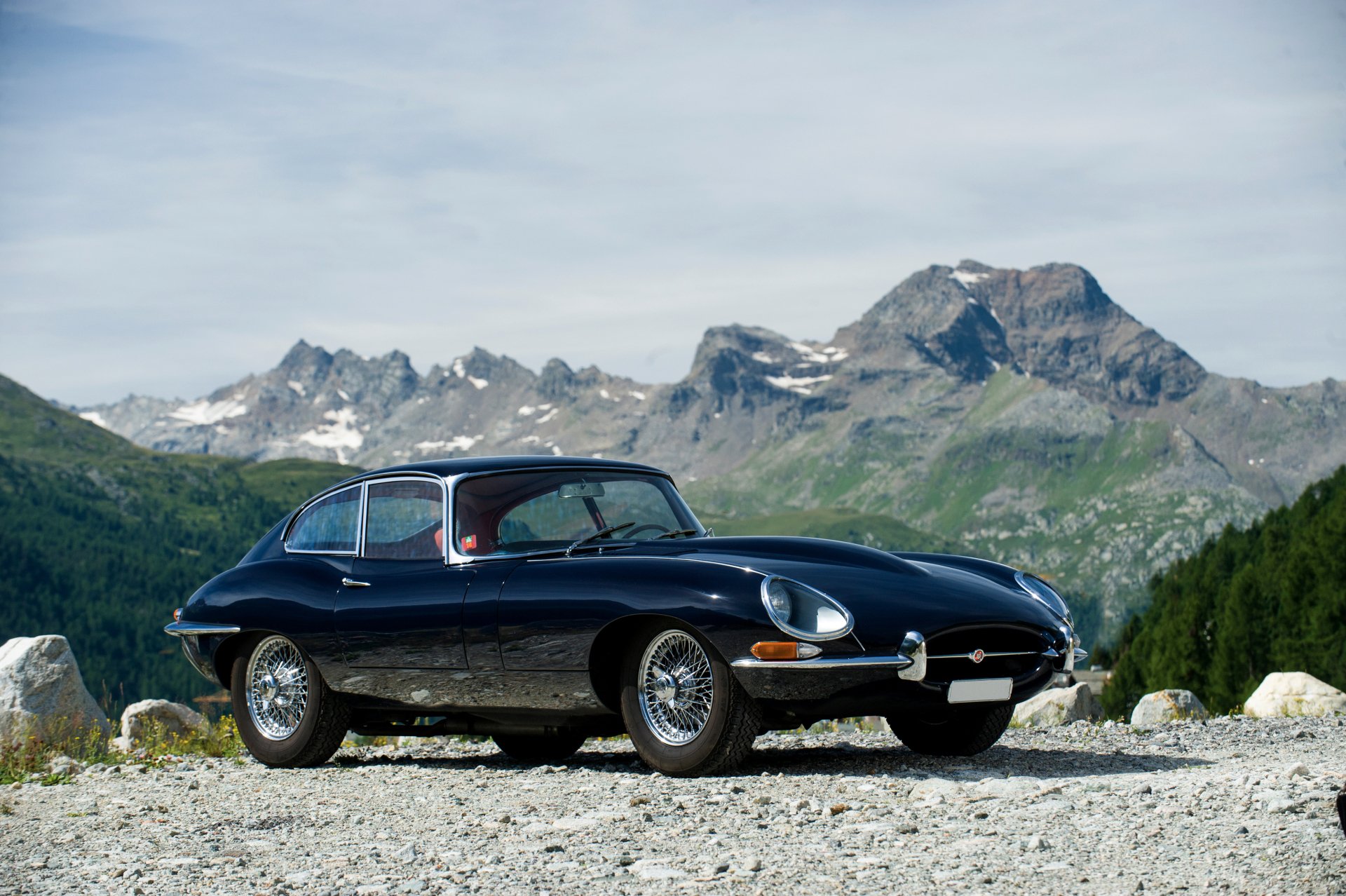 A sleek black Jaguar E-Type supercar parked on a rocky mountain road with towering peaks in the background, captured in stunning 4K Ultra HD.