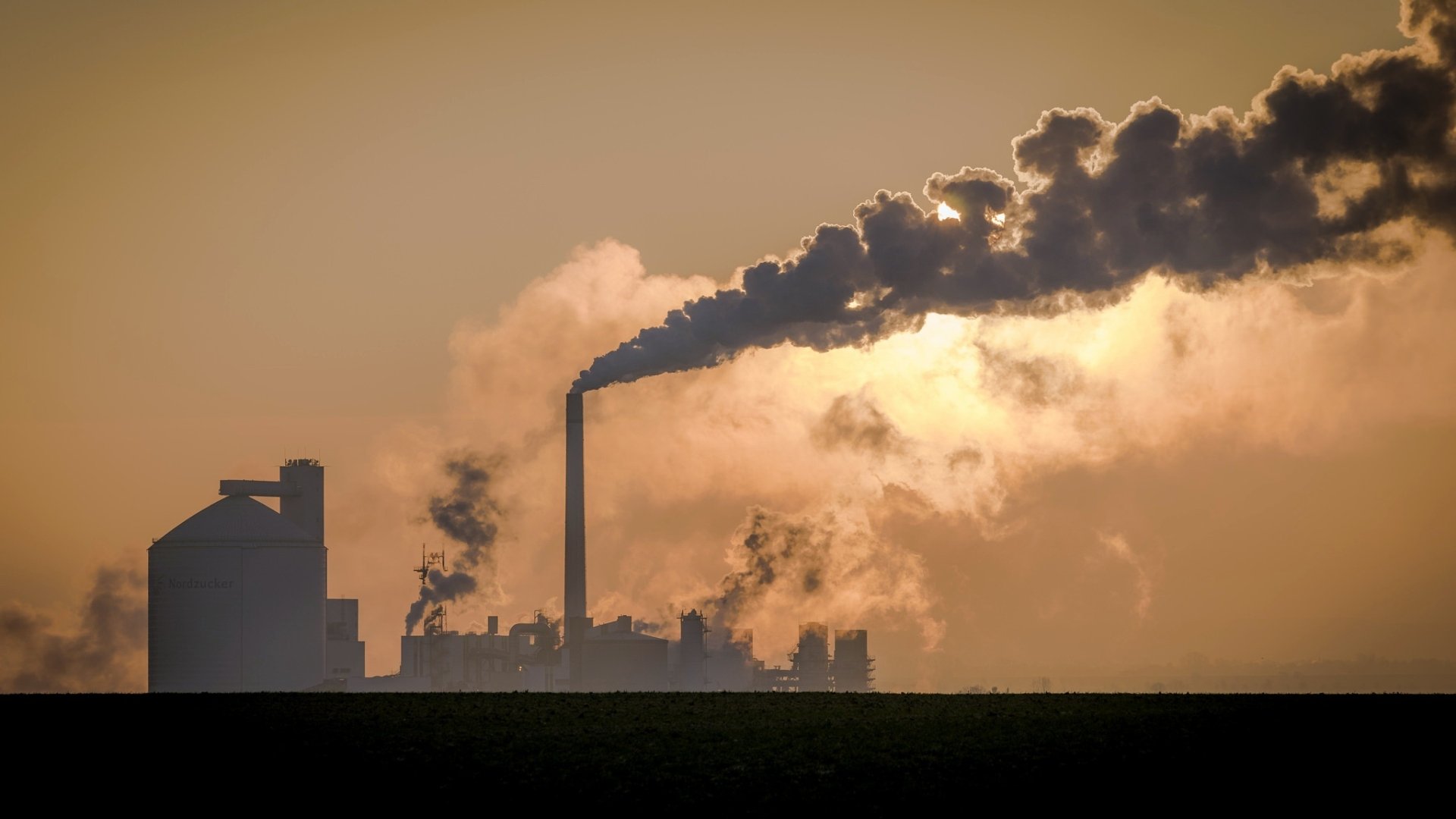 A dramatic sunset silhouettes a factory, with thick smoke billowing from its chimneys, capturing the essence of industrial life against a vibrant sky.