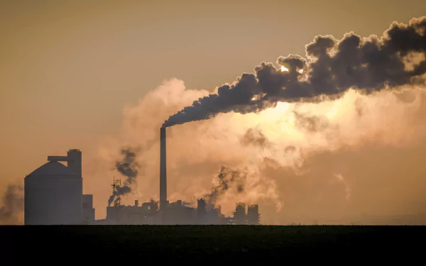 A dramatic sunset silhouettes a factory, with thick smoke billowing from its chimneys, capturing the essence of industrial life against a vibrant sky.