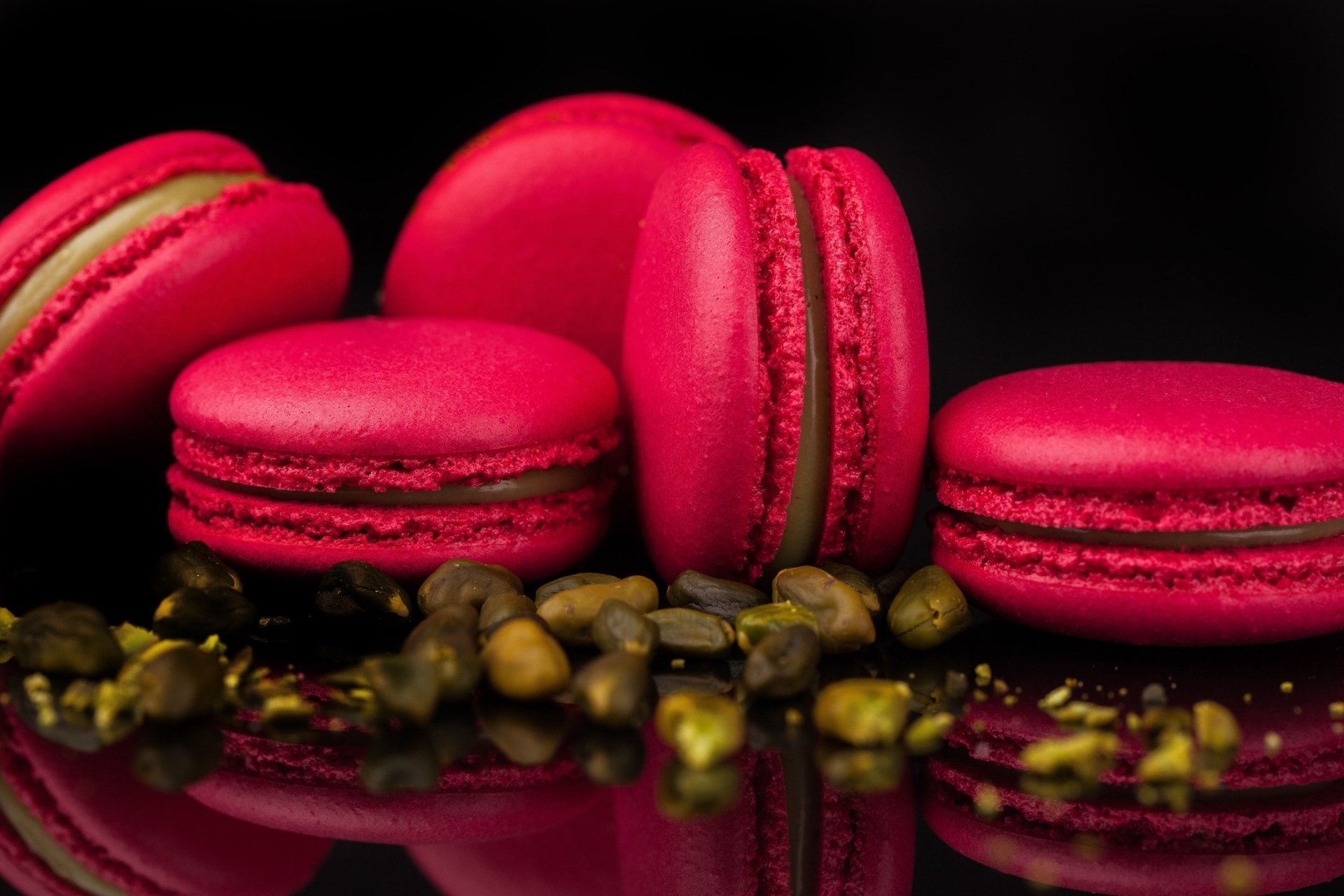Vibrant pink macarons and crushed pistachios on a glossy reflective surface, sweets food close-up — 2K Quad HD PC desktop wallpaper background.