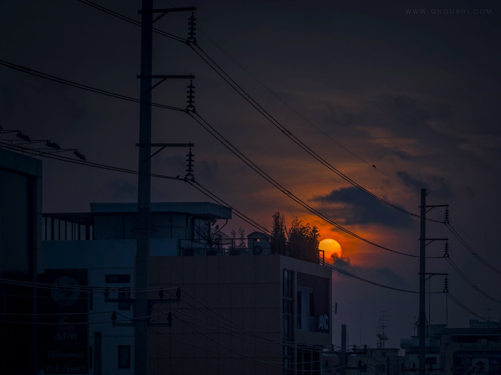HD PC desktop wallpaper: dusk cityscape with building rooftops and power lines silhouetted against a glowing orange sunset — moody photography background.