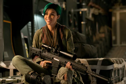 HD desktop wallpaper featuring Ruby Rose from the movie xXx: Return of Xander Cage, sitting with a large firearm, dressed in military attire, smiling confidently in a dimly lit industrial setting.