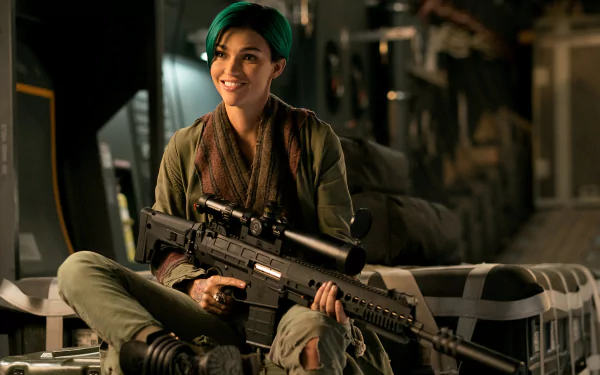 HD desktop wallpaper featuring Ruby Rose from the movie xXx: Return of Xander Cage, sitting with a large firearm, dressed in military attire, smiling confidently in a dimly lit industrial setting.