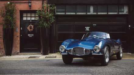 A sleek blue Aston Martin DB2 parked on a street in front of a brick building, captured in this HD PC desktop wallpaper featuring the classic vehicle.