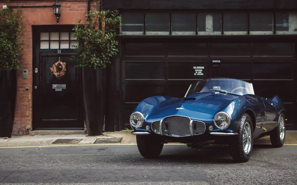 A sleek blue Aston Martin DB2 parked on a street in front of a brick building, captured in this HD PC desktop wallpaper featuring the classic vehicle.