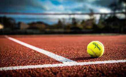 HD desktop wallpaper showing a bright yellow tennis ball on a red clay tennis court under a blue sky, capturing the essence of the sport in vivid detail.