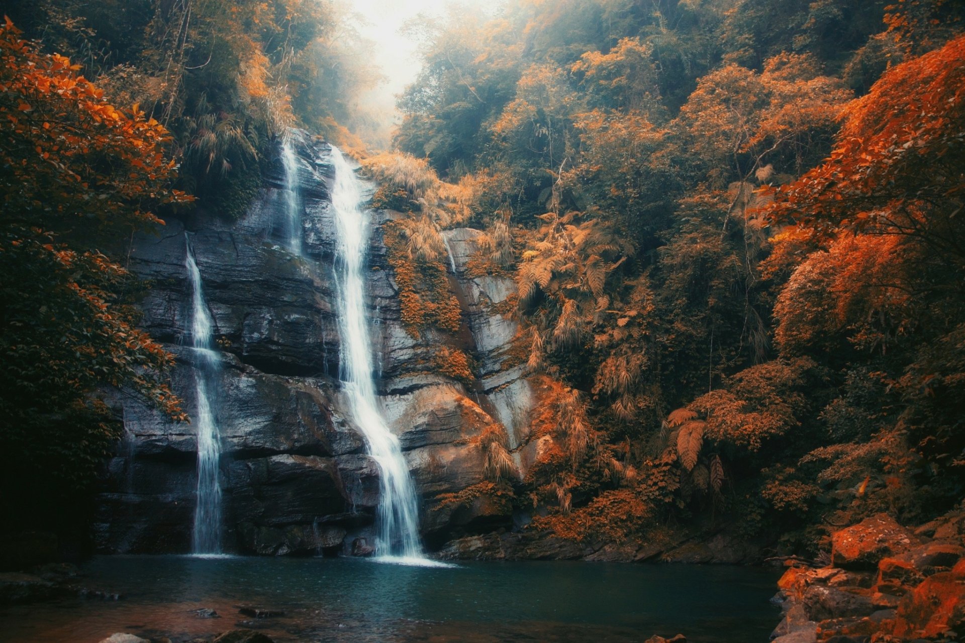 HD desktop wallpaper of a serene waterfall amid fall foliage and gentle fog, showcasing the beauty of nature.