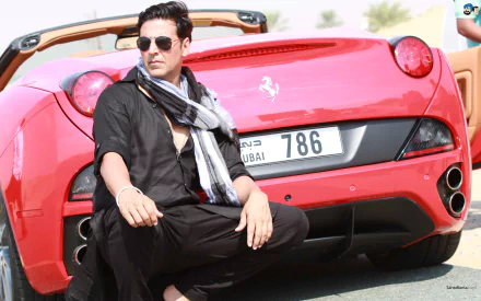 HD desktop wallpaper featuring actor Akshay Kumar, dressed in black with a scarf, sitting on a road by a red sports car with a Dubai license plate.