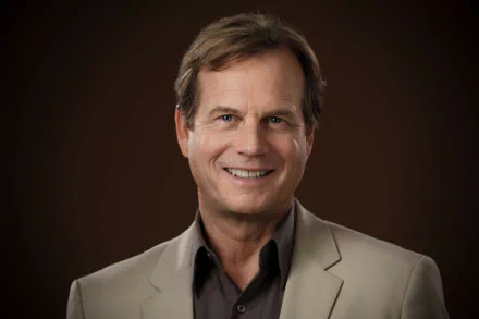 HD desktop wallpaper featuring a smiling person in a beige suit against a brown background.