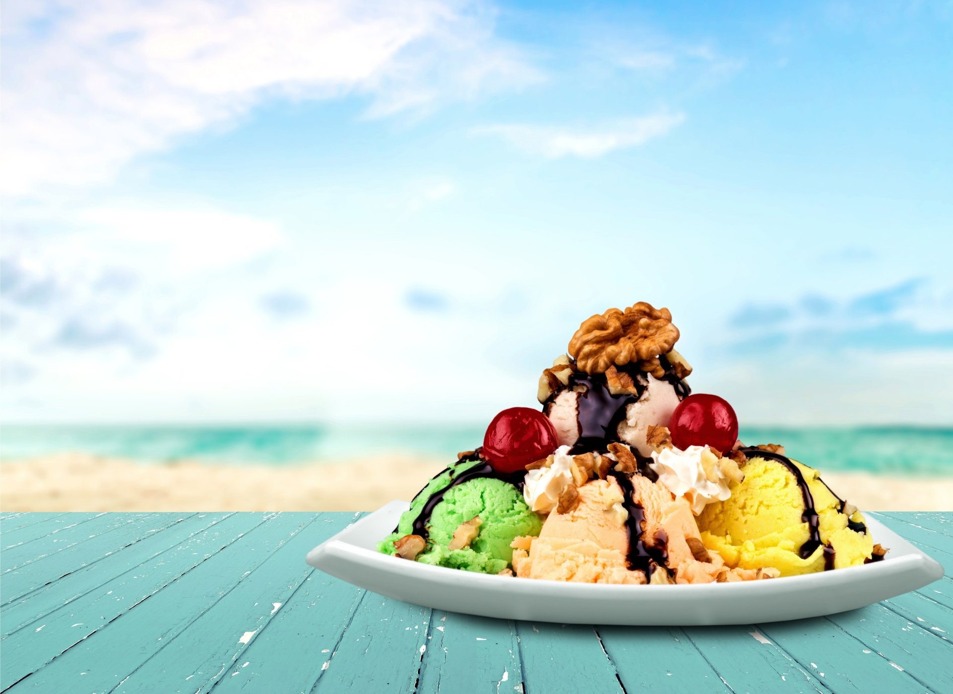 4K Ultra HD desktop wallpaper showing a colorful ice cream sundae with scoops of green, yellow, and white ice cream topped with cherries and chocolate syrup on a beach table.