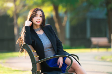 A brunette Asian woman, posed confidently on a park bench, gazes into the camera. The depth of field adds a soft blur to the background. This HD image makes a stylish desktop wallpaper or background.