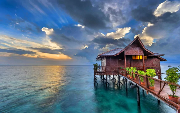HD desktop wallpaper of a tropical overwater bungalow cabin on a man-made wooden pier over turquoise ocean, with dramatic clouds and a glowing horizon.