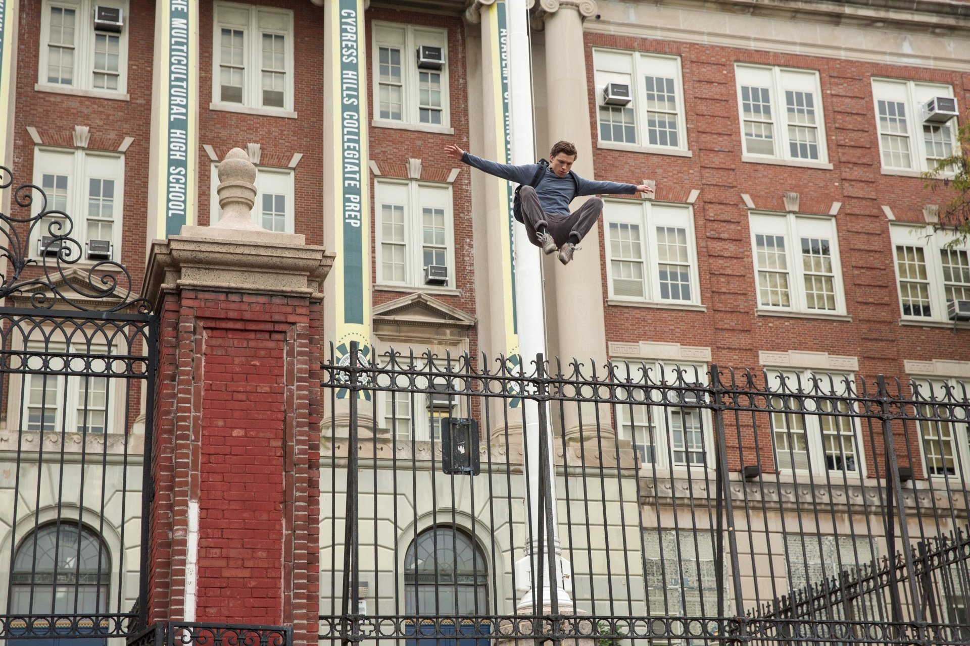 Tom Holland as Peter Parker leaps over a fence in a scene from Spider-Man: Homecoming. HD desktop wallpaper with a school building in the background.