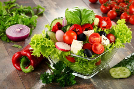 A vibrant still life HD desktop wallpaper featuring a fresh salad with lettuce, tomatoes, pepper, olives, radishes, and feta cheese on a wooden table.