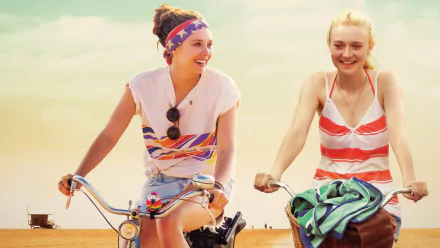 Elizabeth Olsen and Dakota Fanning riding bikes smiling in a scene from the movie Very Good Girls, featured as an HD PC desktop wallpaper and background.