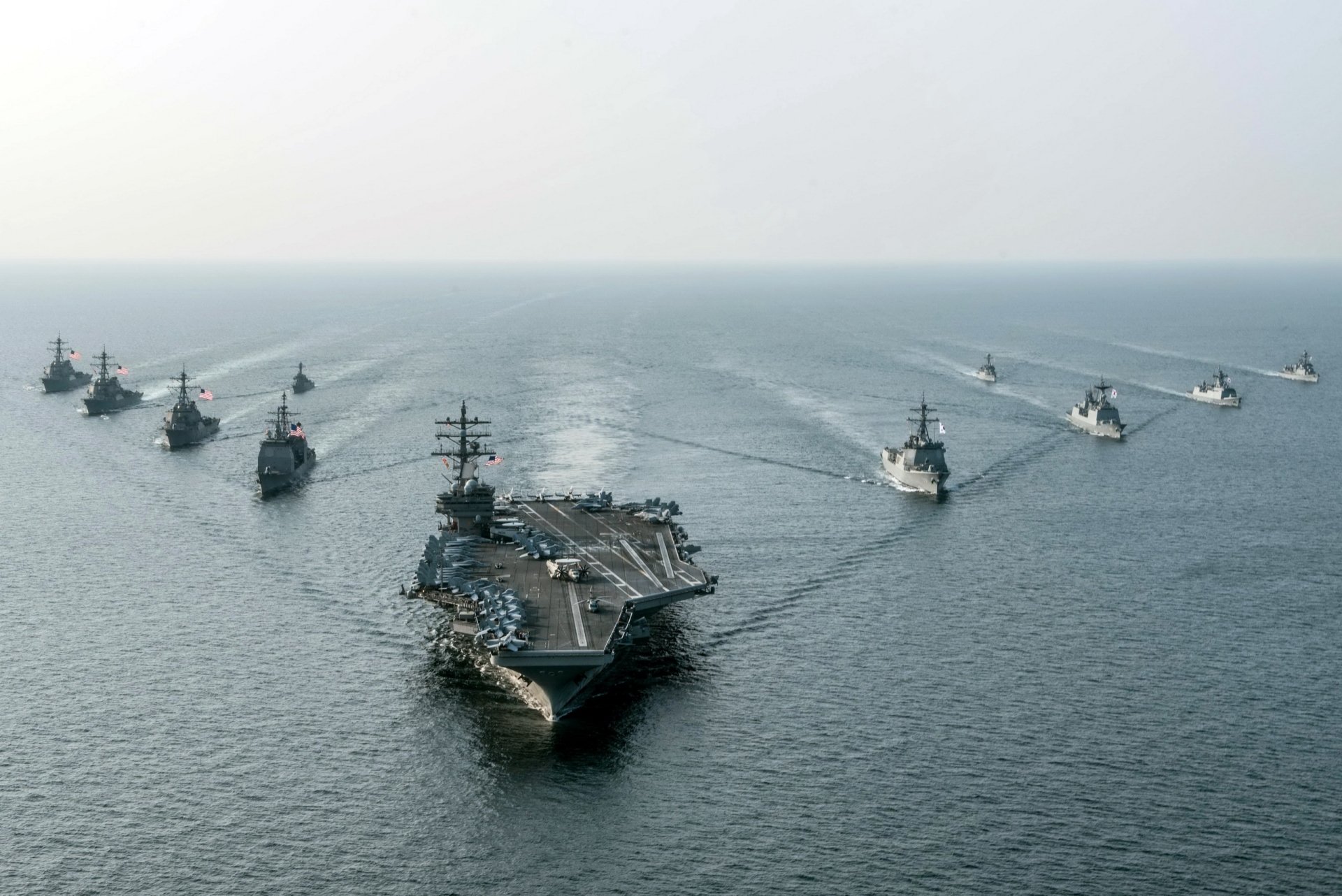 HD desktop wallpaper featuring the USS Ronald Reagan (CVN-76) aircraft carrier leading a naval fleet on the ocean horizon under a clear sky.