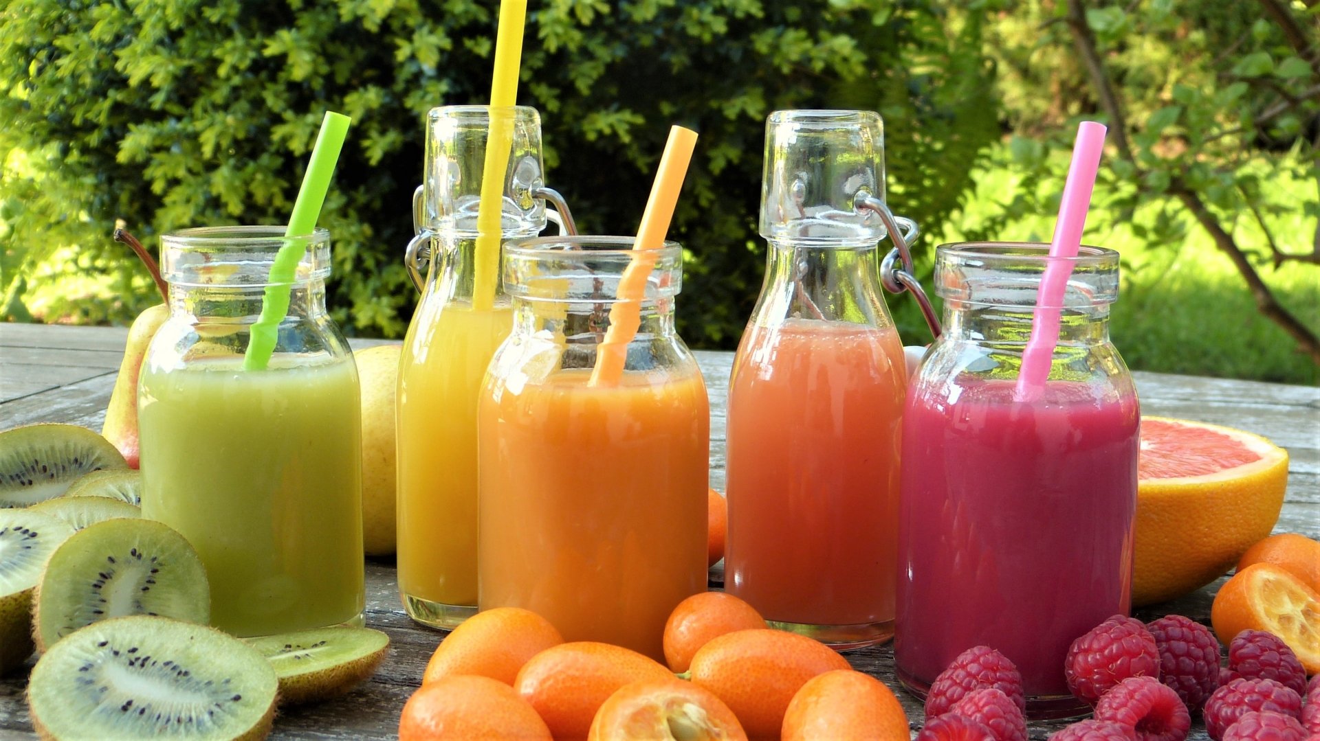 HD desktop wallpaper showing colorful fruit smoothies in glass bottles with straws, surrounded by fresh kiwi, raspberries, and kumquats in an outdoor setting.