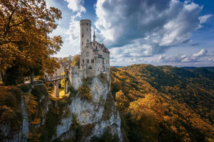 Lichtenstein Castle perched on a cliff, surrounded by vibrant fall forest under a partly cloudy sky, captured as a high-definition desktop wallpaper.