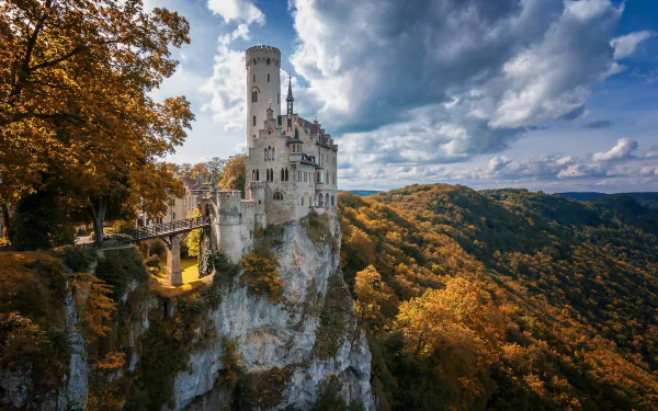 Lichtenstein Castle perched on a cliff, surrounded by vibrant fall forest under a partly cloudy sky, captured as a high-definition desktop wallpaper.
