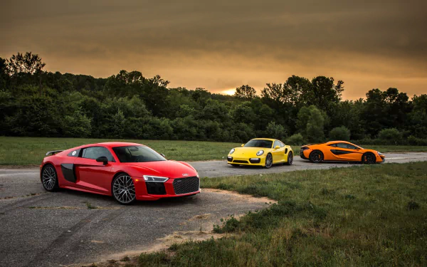 HD desktop wallpaper featuring a red Audi R8 V10, yellow Porsche 911 Turbo, and orange McLaren 570S parked on a scenic road at sunset.