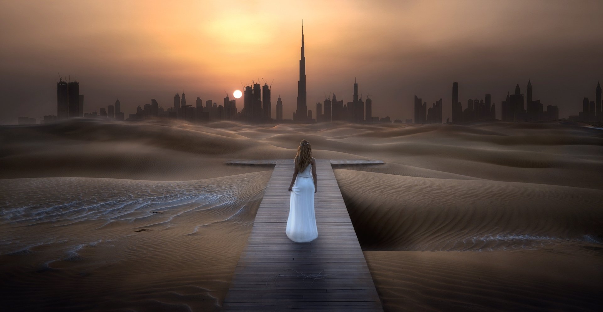A woman in a white dress walks on a wooden path through a desert toward the Burj Khalifa skyscraper at sunset in this surreal HD cityscape wallpaper.