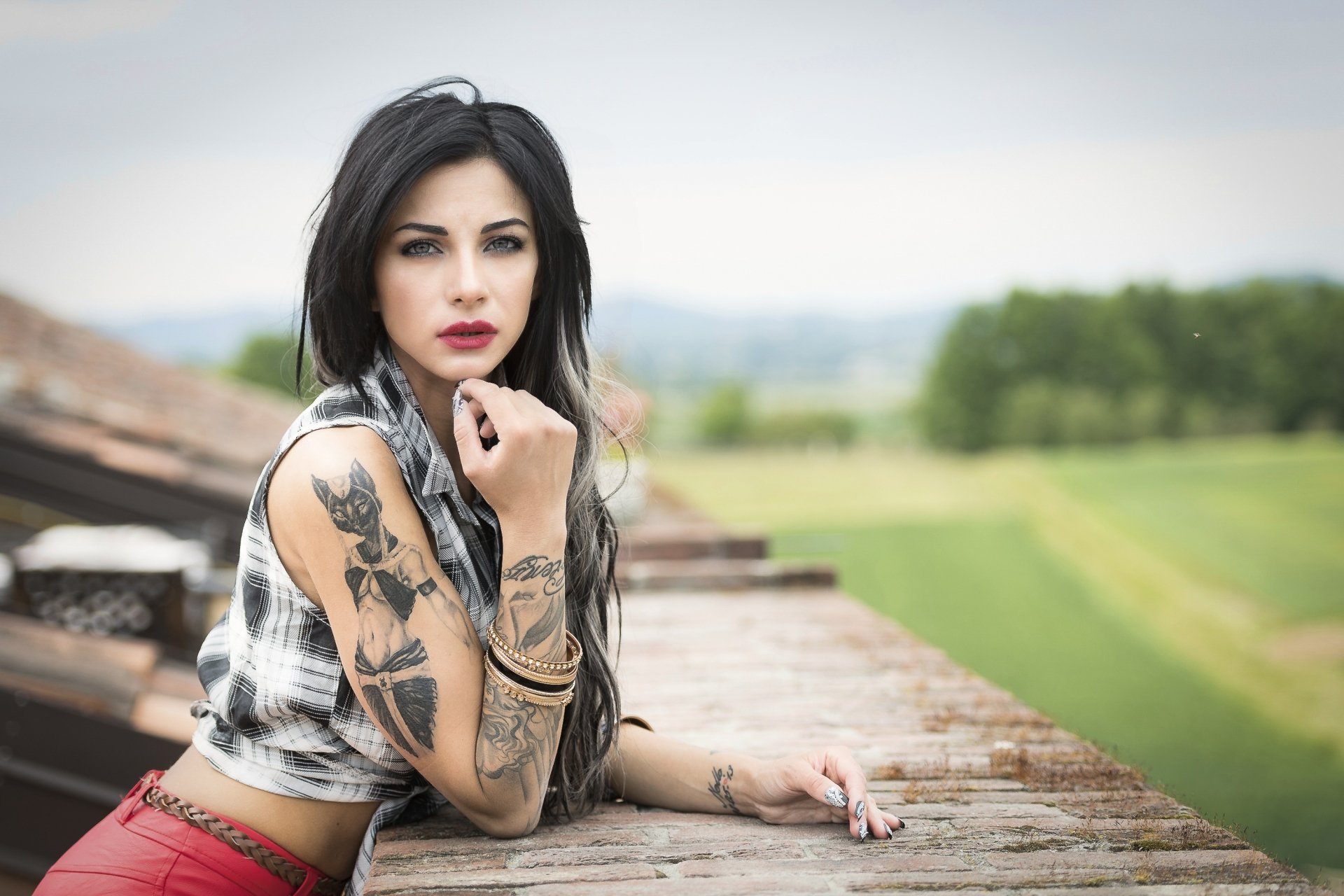 Brunette model with tattoos and lipstick poses outdoors on a rustic wall, captured with shallow depth of field for a striking HD desktop wallpaper background.
