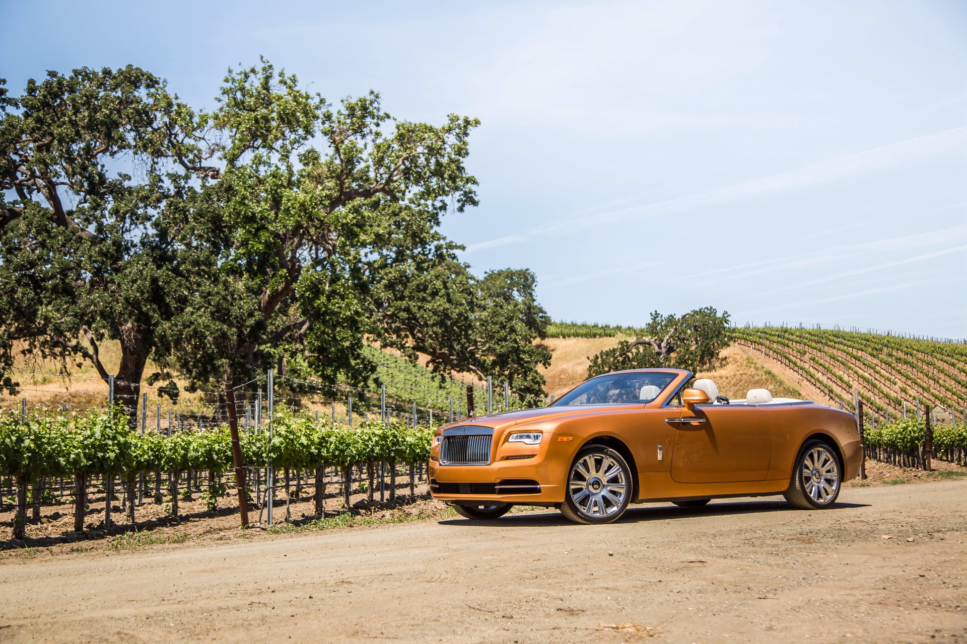 Orange Rolls-Royce Dawn grand tourer convertible parked on a dirt road near vineyards under a clear blue sky, captured in 4K Ultra HD for desktop wallpaper.