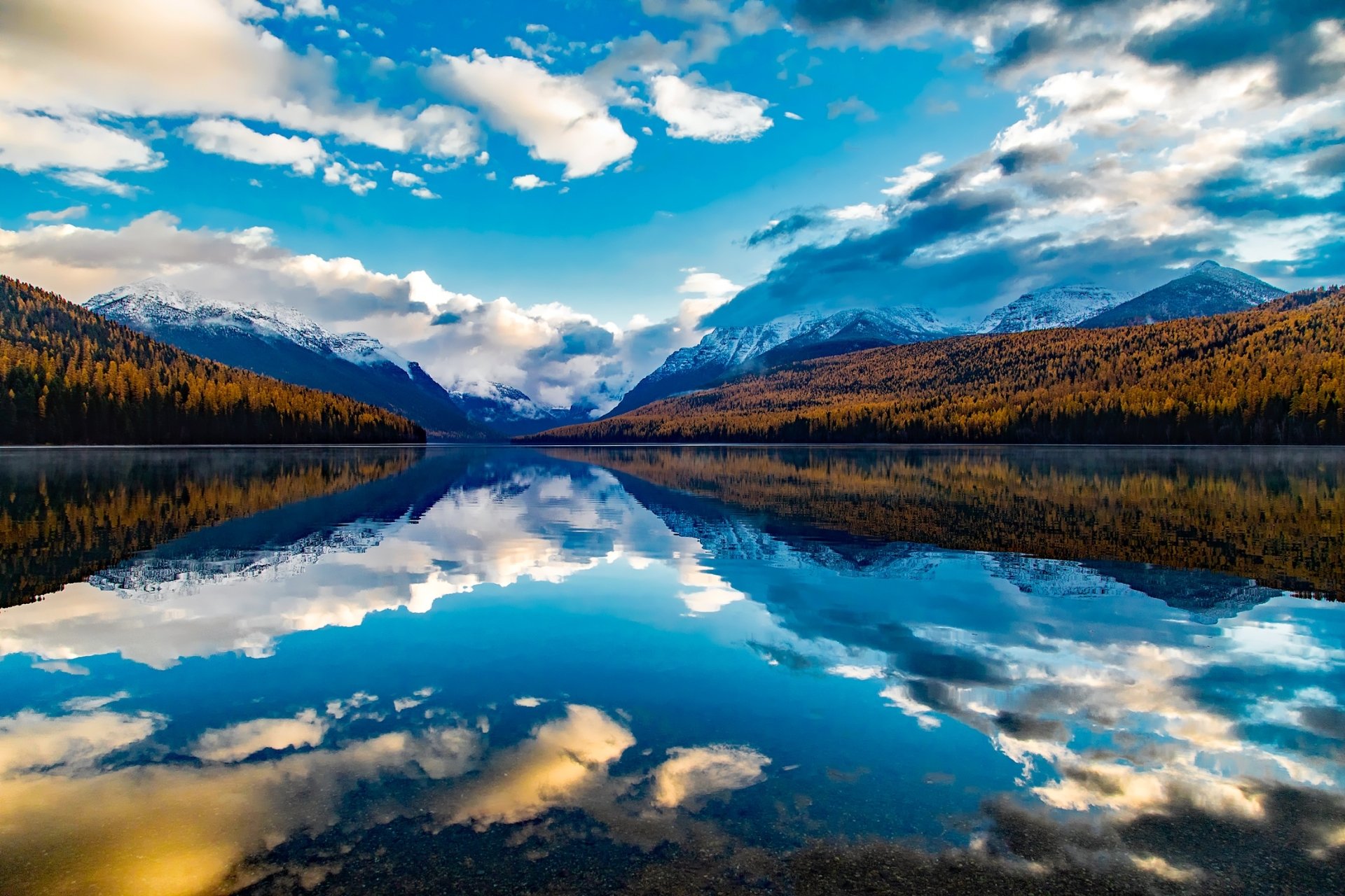 HD desktop wallpaper of Lake McDonald, Glacier National Park: sky and clouds reflected in a glassy lake, framed by snow‑capped mountains and autumn forest.