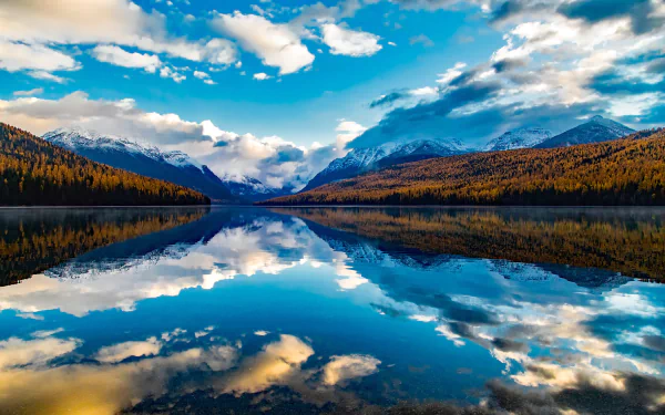 HD desktop wallpaper of Lake McDonald, Glacier National Park: sky and clouds reflected in a glassy lake, framed by snow‑capped mountains and autumn forest.