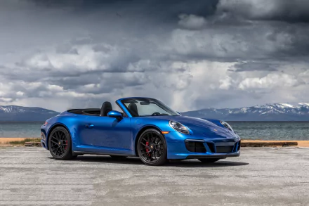 A stunning HD desktop wallpaper featuring a blue Porsche 911 Carrera convertible parked by a serene waterfront, with mountains and dramatic clouds in the background.