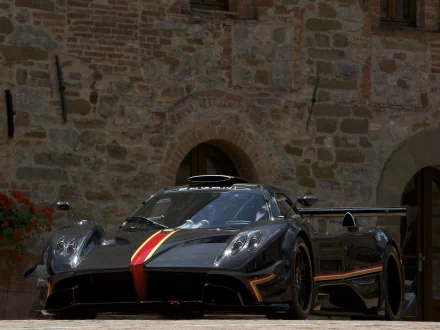 HD PC desktop wallpaper of a black Pagani Zonda R supercar with a red-and-yellow racing stripe, aggressive aero kit and rear wing parked in front of a rustic stone building.