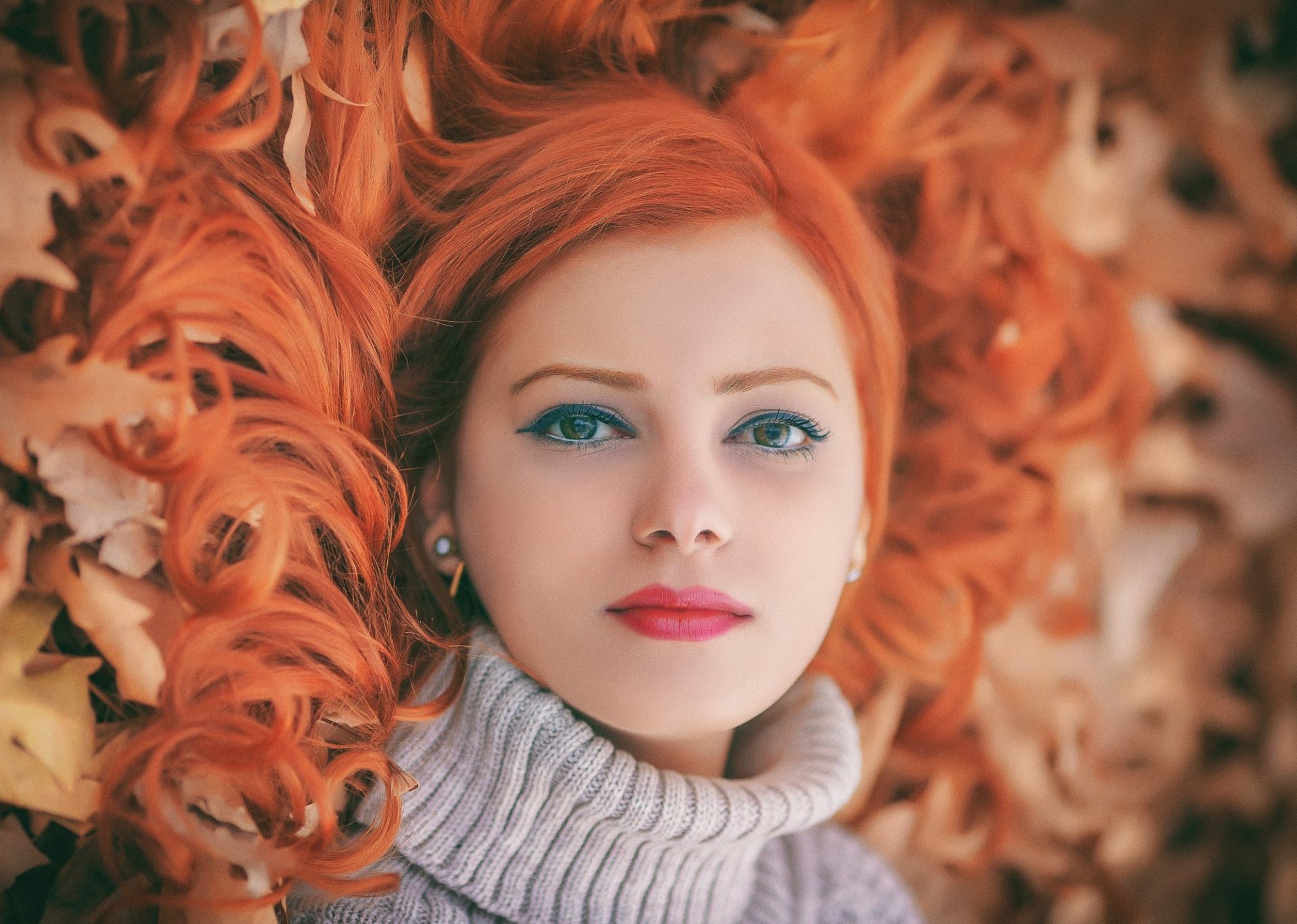 HD desktop wallpaper of a redhead woman with lipstick, gazing confidently, surrounded by autumn leaves, showcasing her vibrant hair and striking facial features.
