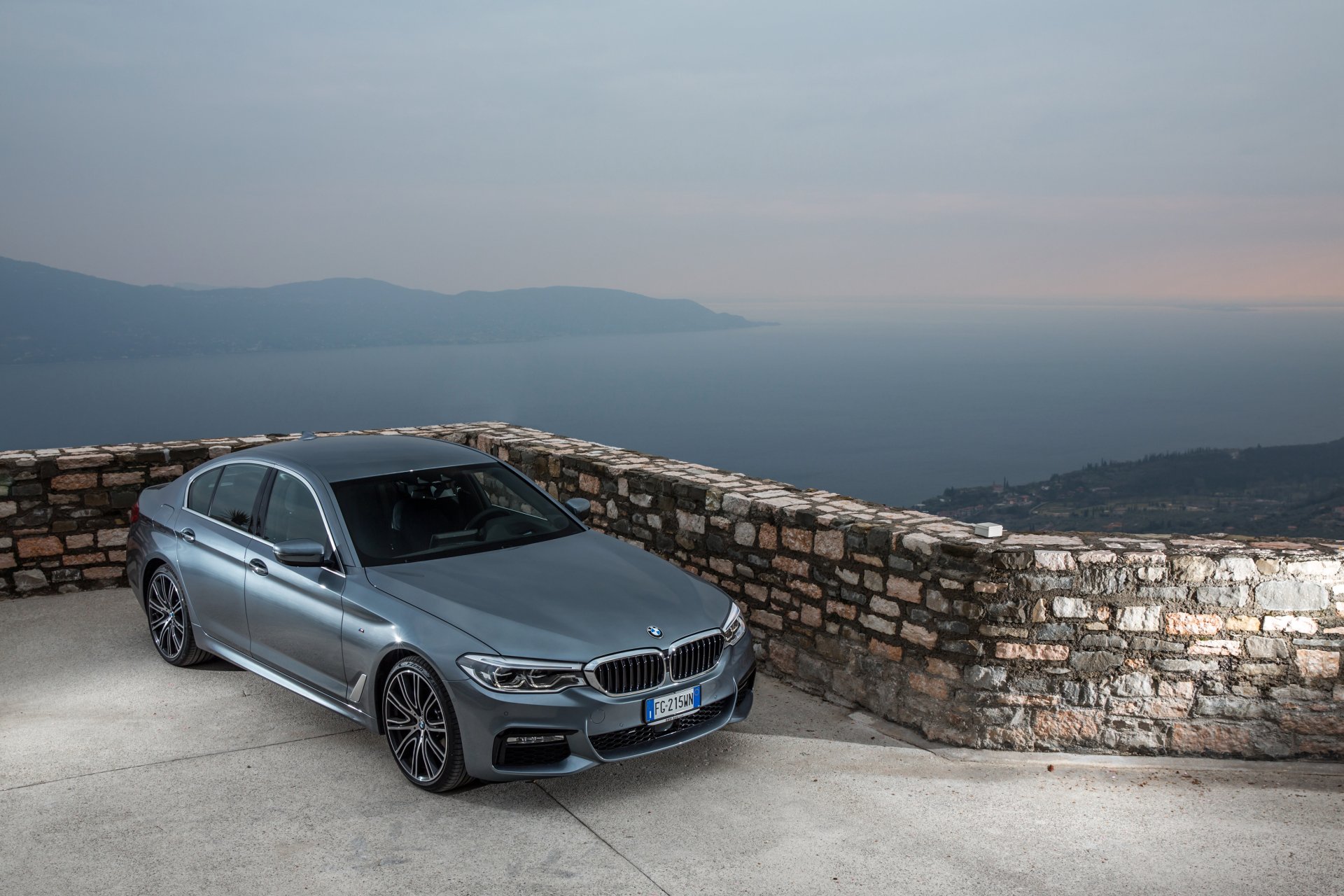 Silver BMW 5 Series parked by a stone wall overlooking a misty landscape, captured in stunning 4K Ultra HD for a PC desktop wallpaper.